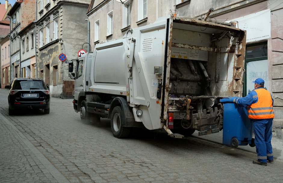 A waste collection worker wearing a red and yellow high-visibility vest is standing next to a large red rubbish truck on a roadside, actively emptying a black plastic bag into the truck's loading comp